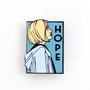 Könnte beinhalten: Rechteckiger Emaille-Pin mit Seitenprofil einer Person mit kurzen blonden Haaren, die eine hellblaue Kapuzenjacke trägt. Das Wort "HOPE" ist vertikal in Schwarz auf hellblauem Hintergrund gedruckt.