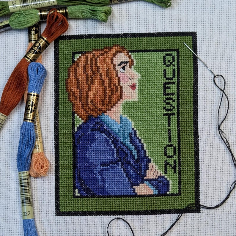 May include: A cross-stitch embroidery of a woman with brown hair wearing a blue jacket. The background is green and the word "QUESTION" is stitched vertically on the right side.