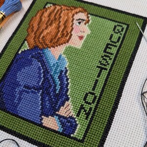 May include: Cross stitch pattern of a woman with brown hair wearing a blue jacket. The background is green and the text "GUEST LIST" is stitched in black.