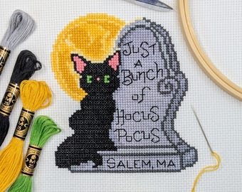 May include: A cross-stitch design featuring a black cat with pink ears, a yellow moon, and a tombstone that reads "Just a Bunch of Hocus Pocus, Salem, MA." Embroidery floss in black, yellow, green, and gray, along with scissors and a hoop, are also visible.