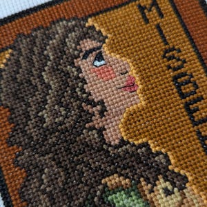 May include: Cross-stitch embroidery of a woman's face with brown hair, wearing a green and brown outfit. The background is a brown frame with the word "Mystery" in black text.