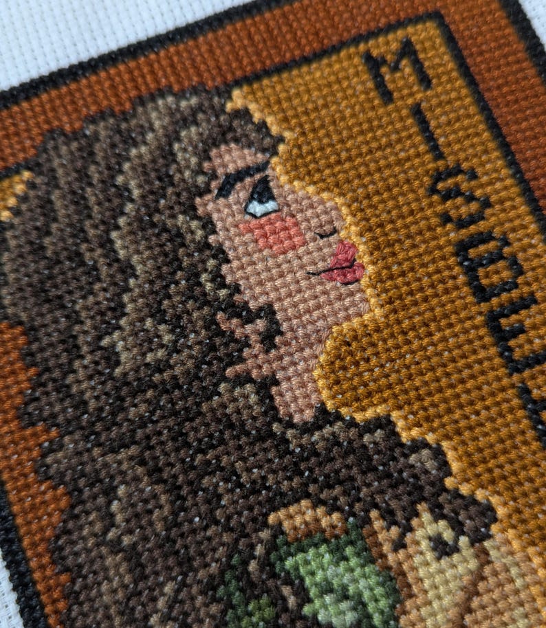 May include: Cross-stitch embroidery of a woman's face with brown hair, wearing a green and brown outfit. The background is a brown frame with the word "Mystery" in black text.