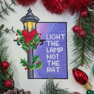 May include: Cross-stitch artwork depicting a lamppost decorated with a red bow and holly, with the text "LIGHT THE LAMP NOT THE RAT" on a purple background. The image is framed by Christmas decorations, including red berries and a decorative light bulb.