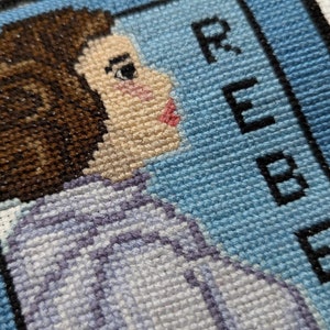 Puede incluir: Bordado de punto de cruz de una mujer con cabello castaño, vistiendo una camisa a rayas azul claro y blanco. El fondo es azul con la palabra "REBEL" bordada en negro.