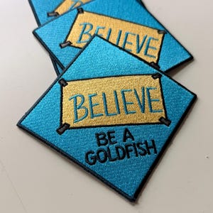 May include: Three square embroidered patches with a bright blue background and a black border. Each patch features a gold rectangular center with the word BELIEVE in blue text. Below the gold rectangle, the text BE A GOLDFISH is embroidered in black.