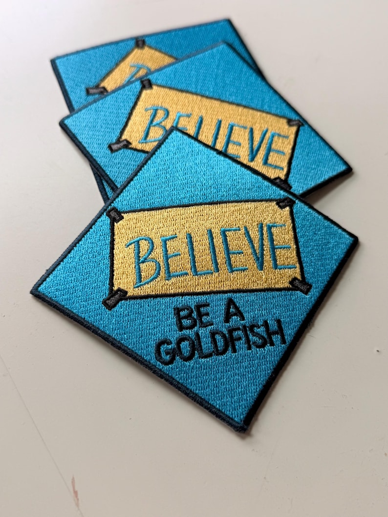 May include: Three square embroidered patches with a bright blue background and a black border. Each patch features a gold rectangular center with the word BELIEVE in blue text. Below the gold rectangle, the text BE A GOLDFISH is embroidered in black.
