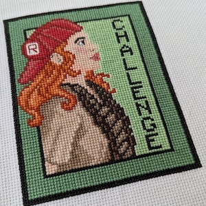 May include: Cross-stitch art featuring a woman with red hair and a red hat, set against a green background. The word "CHALLENGE" is vertically displayed on the right side. The artwork is framed with a black border.