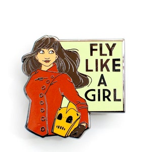 May include: A red and yellow enamel pin featuring a cartoon woman with brown hair wearing a red jacket and holding a yellow robot head. The pin has the text "FLY LIKE A GIRL" on a yellow background.