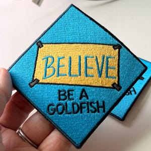 May include: A diamond-shaped embroidered patch with a bright blue background. It features a yellow rectangular center with the word BELIEVE in blue embroidery, and the text BE A GOLDFISH in black embroidery below the yellow rectangle.