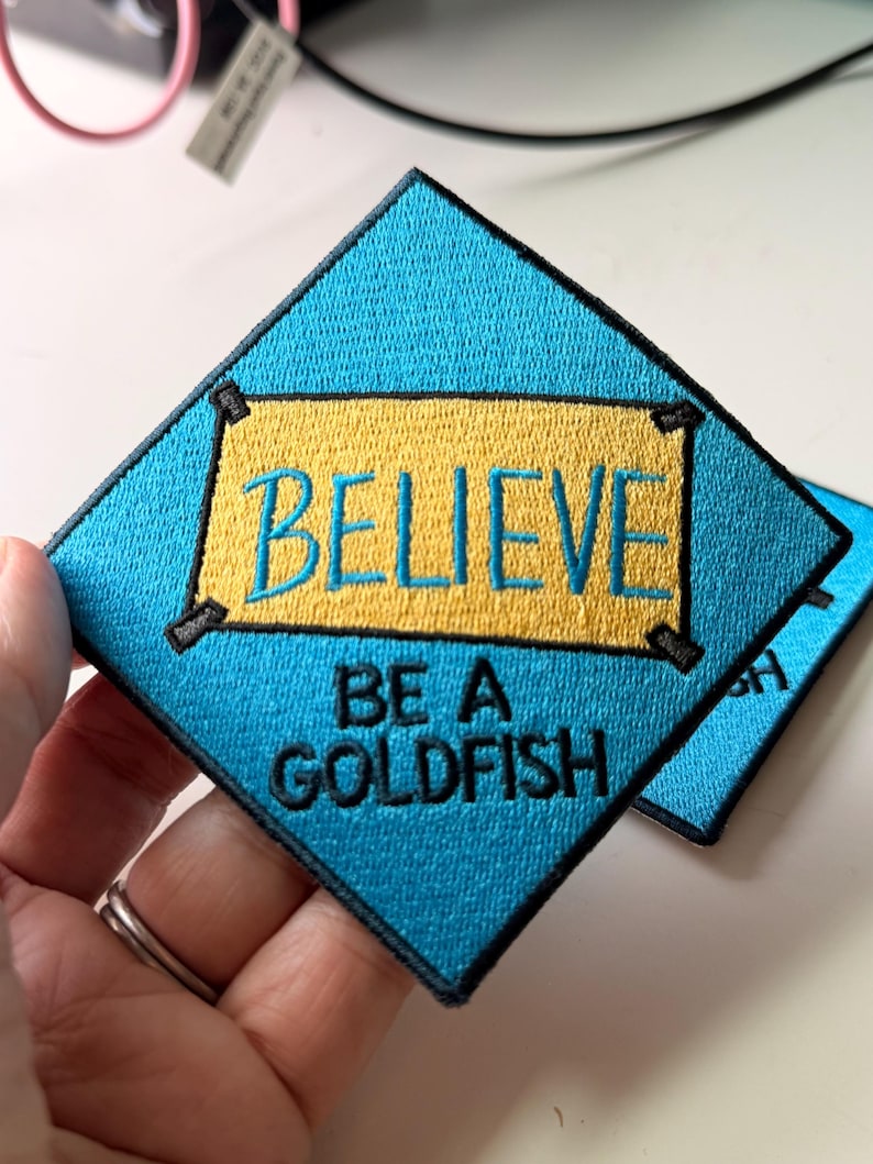 May include: A diamond-shaped embroidered patch with a bright blue background. It features a yellow rectangular center with the word BELIEVE in blue embroidery, and the text BE A GOLDFISH in black embroidery below the yellow rectangle.