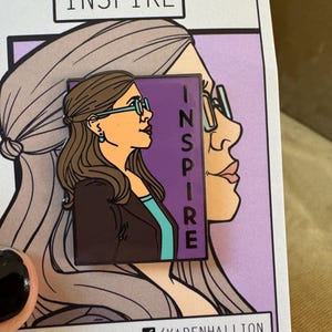 Op de afbeelding: Een paarse en turquoise emaille pin met een cartoonillustratie van een vrouw met een bril en het woord "INSPIRE" verticaal geschreven. De pin is op een witte kaart met de tekst "INSPIRE" en de naam van de kunstenaar en zijn social media-handvatten.