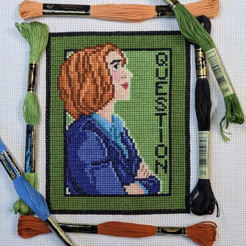 May include: Cross stitch pattern of a woman with brown hair wearing a blue jacket. The background is green and the word "QUESTION" is stitched vertically on the right side of the image.