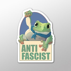May include: A sticker depicting a cartoon frog wearing a blue bandana, raising a fist. The frog is holding a sign with the words "ANTI FASCIST" in green. The background is a blue oval shape.