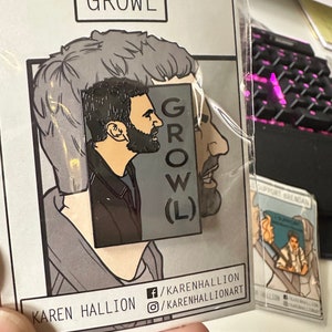 May include: A pin badge with a side profile of a man with a beard, set against a gray background with the word "GROWL" in a rectangular frame. The badge is in a clear plastic sleeve. The artist's name and social media handles are visible.