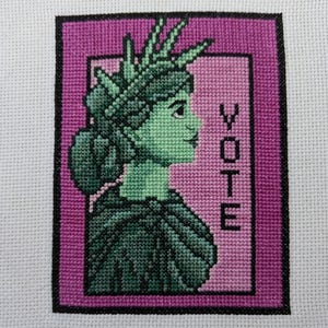 May include: Cross-stitch embroidery of a woman with a crown, in profile, with the word "VOTE" in the background. The woman is green and the background is pink.