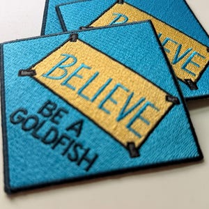 May include: Two square embroidered patches with a bright blue background and a yellow rectangular center. The yellow section features the word BELIEVE in blue thread, and the blue area below features the black text BE A GOLDFISH.