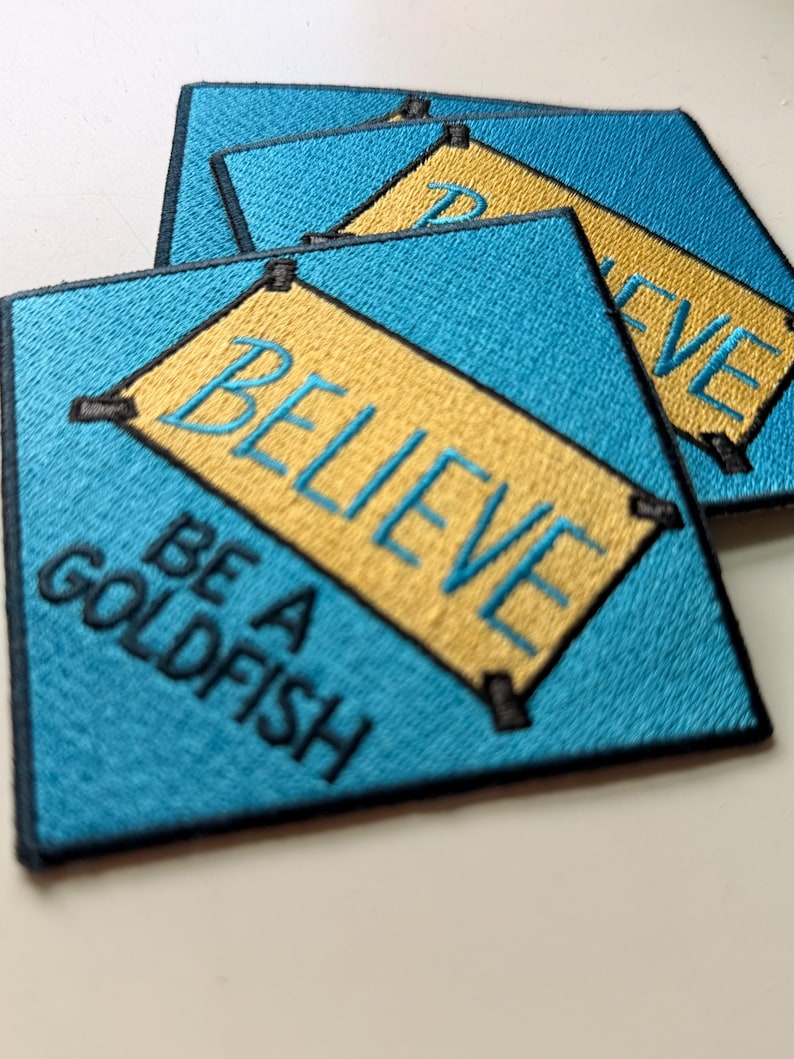 May include: Two square embroidered patches with a bright blue background and a yellow rectangular center. The yellow section features the word BELIEVE in blue thread, and the blue area below features the black text BE A GOLDFISH.