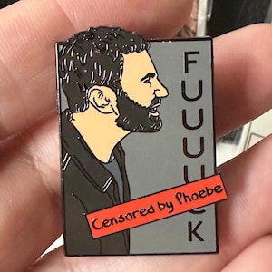May include: A rectangular enamel pin featuring a profile of a man with a beard. The pin has the word "FUCK" vertically on the right side, with a red banner that says "Censored by Phoebe".