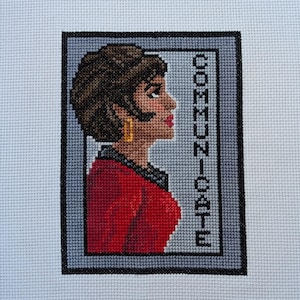 Peut inclure: Broderie au point de croix d'une femme aux cheveux bruns portant une veste rouge et des boucles d'oreilles dorées. Le mot "COMMUNICATE" est brodé verticalement sur le côté droit de l'image.