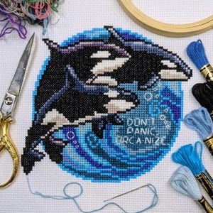 May include: A completed cross-stitch featuring two orcas in a blue ocean scene. The design includes the text "Don't Panic Orca-nize". Embroidery floss, scissors, and a needle are visible.