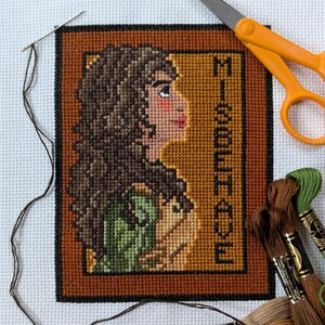 May include: Cross stitch pattern of a woman with brown hair and a green dress. The pattern is framed in a brown frame with the text "Misbehave" in the corner.