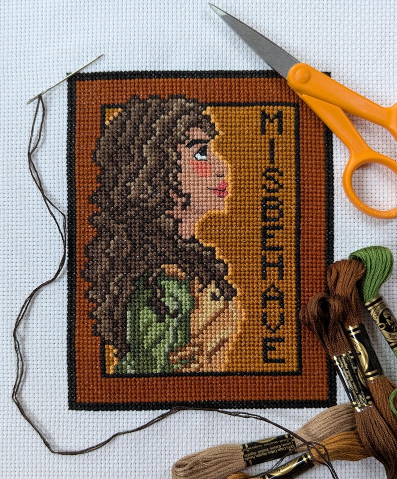 May include: Cross stitch pattern of a woman with brown hair and a green dress. The pattern is framed in a brown frame with the text "Misbehave" in the corner.
