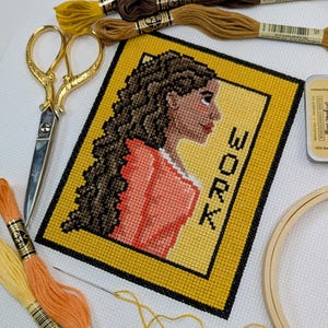 May include: A cross-stitch artwork featuring a woman's profile with long, curly hair, wearing a red top, and the word "WORK" in black letters. The artwork is framed in yellow, with gold scissors, embroidery floss, and a needle nearby.