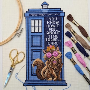 May include: A cross-stitch project on white fabric featuring a blue police box with the text "YOU KNOW HOW I FEEL ABOUT TIME TRAVEL, CARL" and a brown cat wearing pink glasses and a crown, with embroidery scissors, a hoop, and thread.