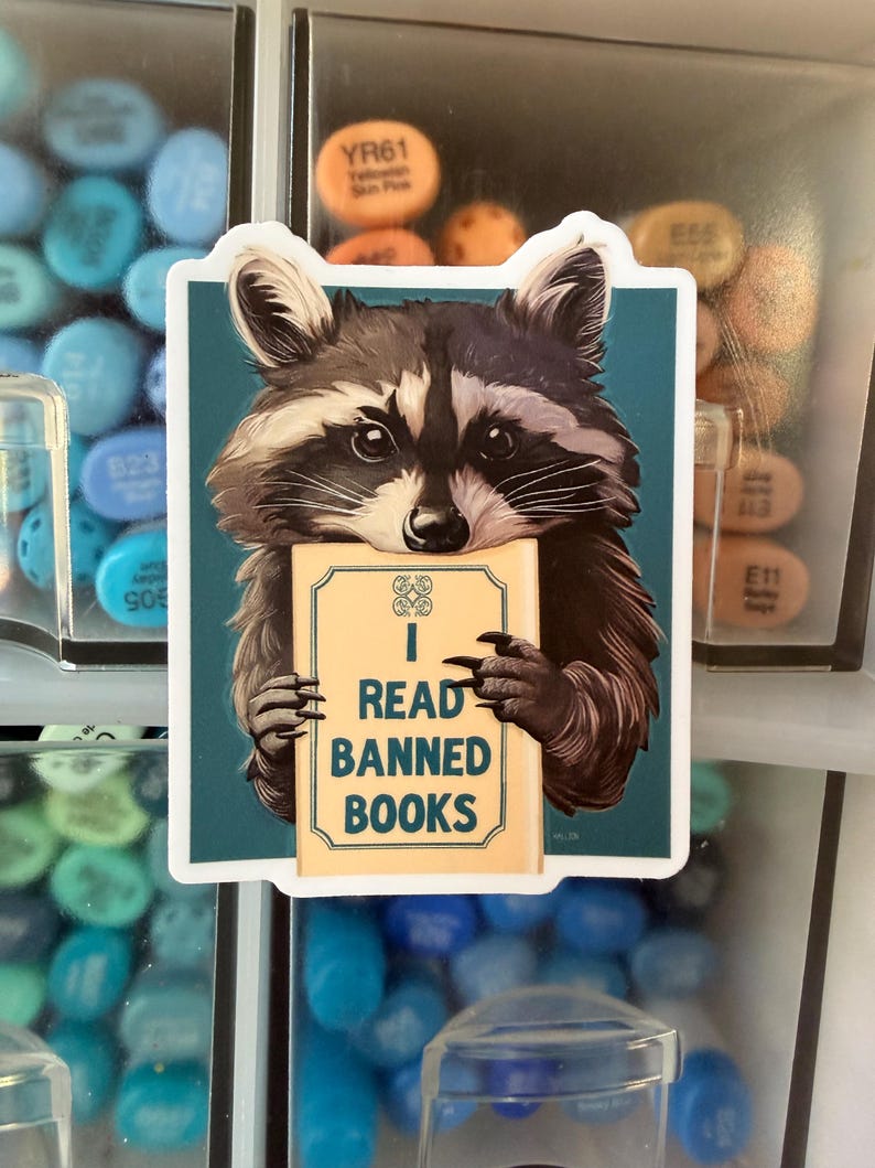 May include: A rectangular sticker featuring a detailed illustration of a raccoon with black and grey fur holding a light tan book. The book cover displays the text I READ BANNED BOOKS in dark blue letters. The background is a solid teal color.