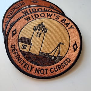 Op de afbeelding: Twee ronde geborduurde patches met een vuurtoren en een zeilboot op een beige achtergrond. De rand is koperkleurig met zwarte tekst WIDOW'S BAY en DEFINITELY NOT CURSED, gescheiden door kleine zwarte ruitvormen aan beide zijden.