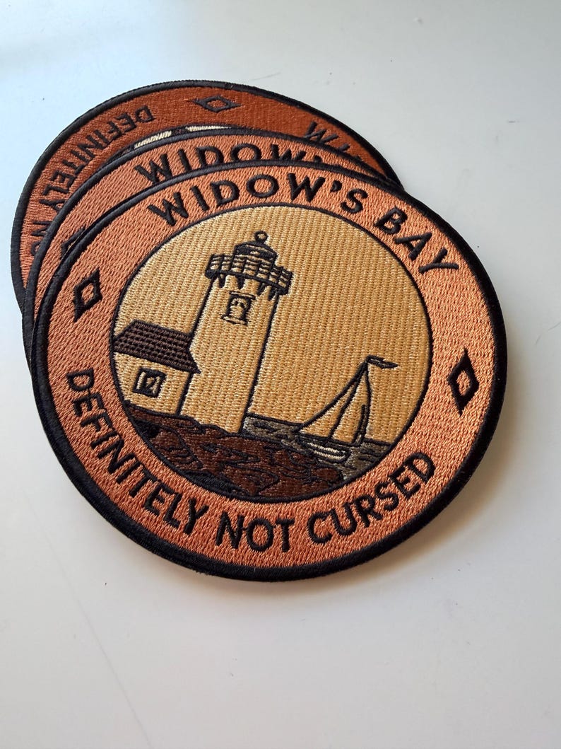 Op de afbeelding: Twee ronde geborduurde patches met een vuurtoren en een zeilboot op een beige achtergrond. De rand is koperkleurig met zwarte tekst WIDOW'S BAY en DEFINITELY NOT CURSED, gescheiden door kleine zwarte ruitvormen aan beide zijden.