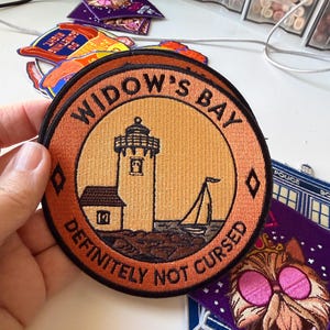 Op de afbeelding: Een ronde geborduurde patch met een vuurtoren en een zeilboot op een lichtoranje achtergrond. De zwarte rand bevat de tekst WIDOW'S BAY bovenaan en DEFINITELY NOT CURSED onderaan, gescheiden door twee kleine zwarte ruitvormen.
