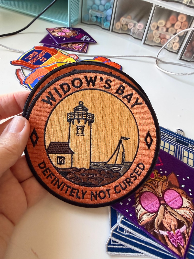 Op de afbeelding: Een ronde geborduurde patch met een vuurtoren en een zeilboot op een lichtoranje achtergrond. De zwarte rand bevat de tekst WIDOW'S BAY bovenaan en DEFINITELY NOT CURSED onderaan, gescheiden door twee kleine zwarte ruitvormen.