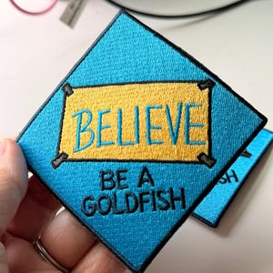 May include: A diamond-shaped embroidered patch with a bright blue background and a central yellow rectangular banner. The text BELIEVE is stitched in blue on the yellow banner, and the text BE A GOLDFISH is stitched in black below it.