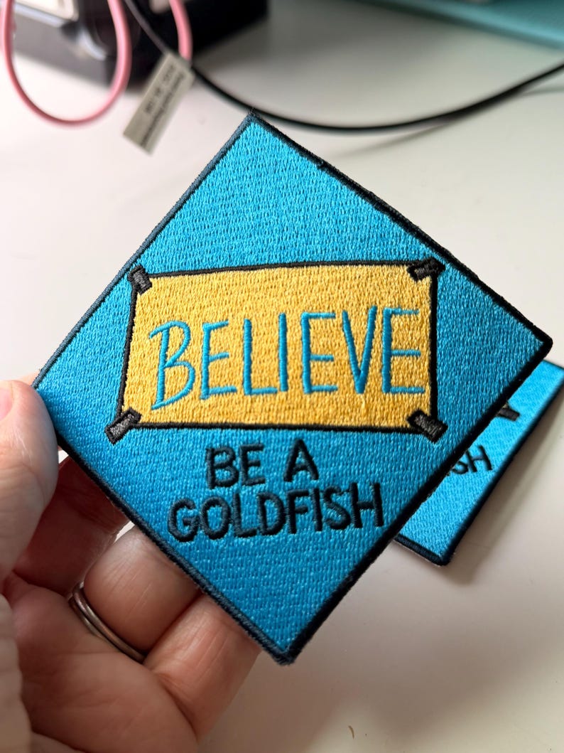 May include: A diamond-shaped embroidered patch with a bright blue background and a central yellow rectangular banner. The text BELIEVE is stitched in blue on the yellow banner, and the text BE A GOLDFISH is stitched in black below it.