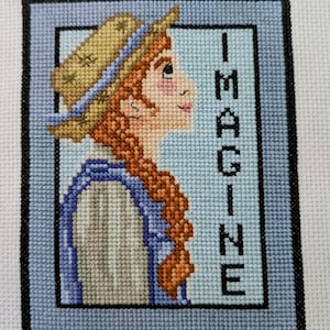 May include: Cross-stitch embroidery of a girl with red hair wearing a straw hat and a blue and white striped shirt. The background is blue and the word "IMAGINE" is stitched in black.
