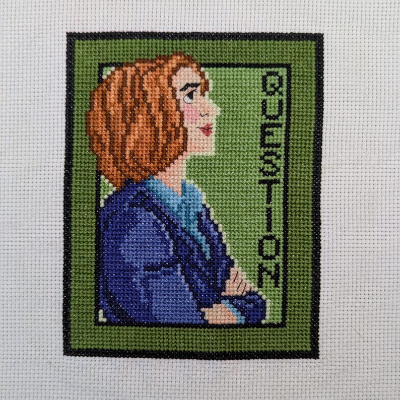May include: Cross-stitch embroidery of a woman with brown hair wearing a blue jacket. The background is green with the word "QUESTION" in white.