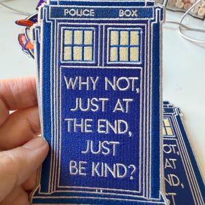 May include: A blue embroidered patch shaped like a police box with white text that reads: WHY NOT, JUST AT THE END, JUST BE KIND? The patch features a white window grid at the top and a small white light fixture at the very peak of the structure.