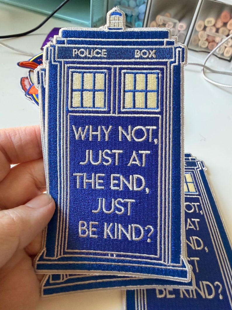May include: A blue embroidered patch shaped like a police box with white text that reads: WHY NOT, JUST AT THE END, JUST BE KIND? The patch features a white window grid at the top and a small white light fixture at the very peak of the structure.