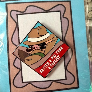 May include: A diamond-shaped enamel pin featuring a cartoon pig wearing a tan fedora and trench coat. The pin has a red banner at the bottom with the white text "BETTER A PIG THAN A FASCIST". The backing card displays the text "BETTER A PIG".
