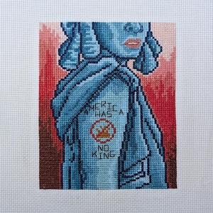May include: Cross-stitch artwork showing a blue figure with the words "America Has No King." The figure is set against a red and brown backdrop. The text is white, with a red circle around a gold crown.
