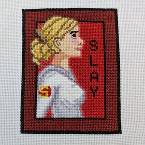 May include: Cross-stitch embroidery of a woman with blonde hair in a white shirt with a red "S" on the sleeve. The background is red with the word "SLAY" in white.