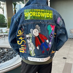 May include: A blue denim jacket with colorful painted designs. The jacket features a skeleton figure with dollar bills, a red and black figure with an X on its forehead, and a blue and purple figure with a bandage. The jacket also has a yellow patch that says "WORLDWIDE" and the text "Hustle" on the left side. The jacket has a white tag with the coordinates "34.0522° N, 118.2437° W".