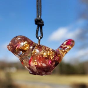 May include: A small bird-shaped pendant made of clear resin with embedded gold leaf and dried rose petals. The pendant is hanging from a black cord.
