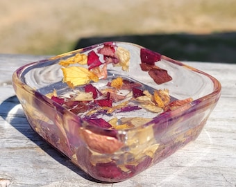 May include: A clear square dish filled with dried rose petals and leaves. The petals are a mix of red, pink, and yellow.