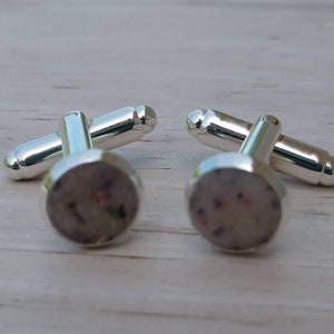 May include: A pair of silver cufflinks with a round, white, and purple design. The cufflinks have a polished silver finish.