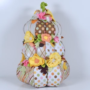 May include: A decorative, stacked pumpkin with a variety of patterned fabrics, including polka dots, plaid, and argyle. The pumpkin is adorned with colorful felt flowers and leaves in shades of yellow, pink, and orange, creating a festive autumn theme.