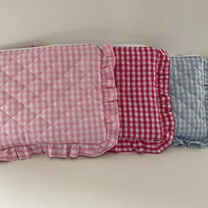 Può includere: Tre pochette in tessuto trapuntato con motivi a quadretti rosa, rossi e blu. Ogni pochette ha un bordo a balze e un motivo trapuntato a rombi. Le pochette sono disposte una accanto all'altra.