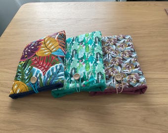 Puede incluir: Tres bolsas de tela con diferentes patrones. La bolsa de la izquierda tiene un patrón de hojas tropicales en tonos de verde, azul, naranja y rojo. La bolsa del medio tiene un patrón de cactus verde y blanco. La bolsa de la derecha tiene un patrón floral en tonos de marrón, rosa y blanco. Cada bolsa tiene un cierre de botón de madera.