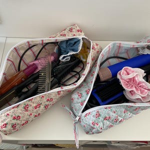 Puede incluir: Dos neceseres con estampado floral, uno con rayas rojas y blancas y otro con rayas azules y blancas. Las bolsas están llenas de herramientas de peinado, incluyendo una plancha, un rizador y cepillos.